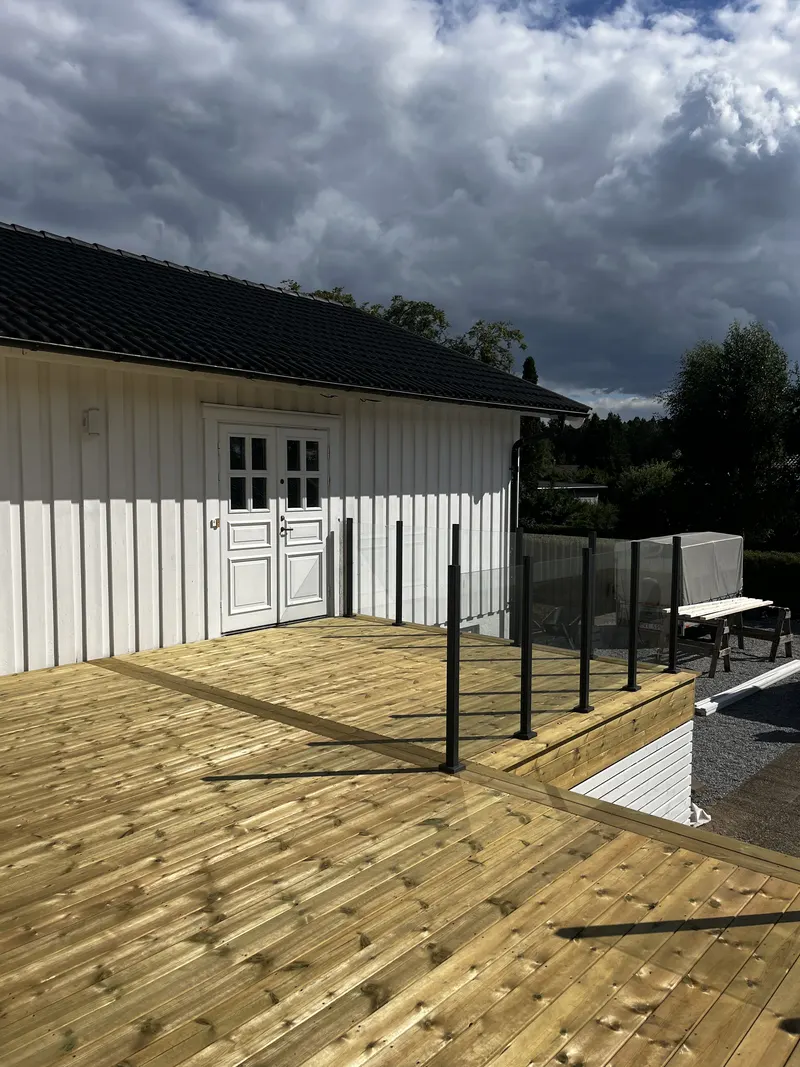 Altan i trä med svarta glasräckesposter och vita inramade fönster mot trädgården